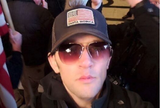 Man wearing sunglasses and a baseball cap with an American flag, taking a selfie in a crowded indoor setting.
