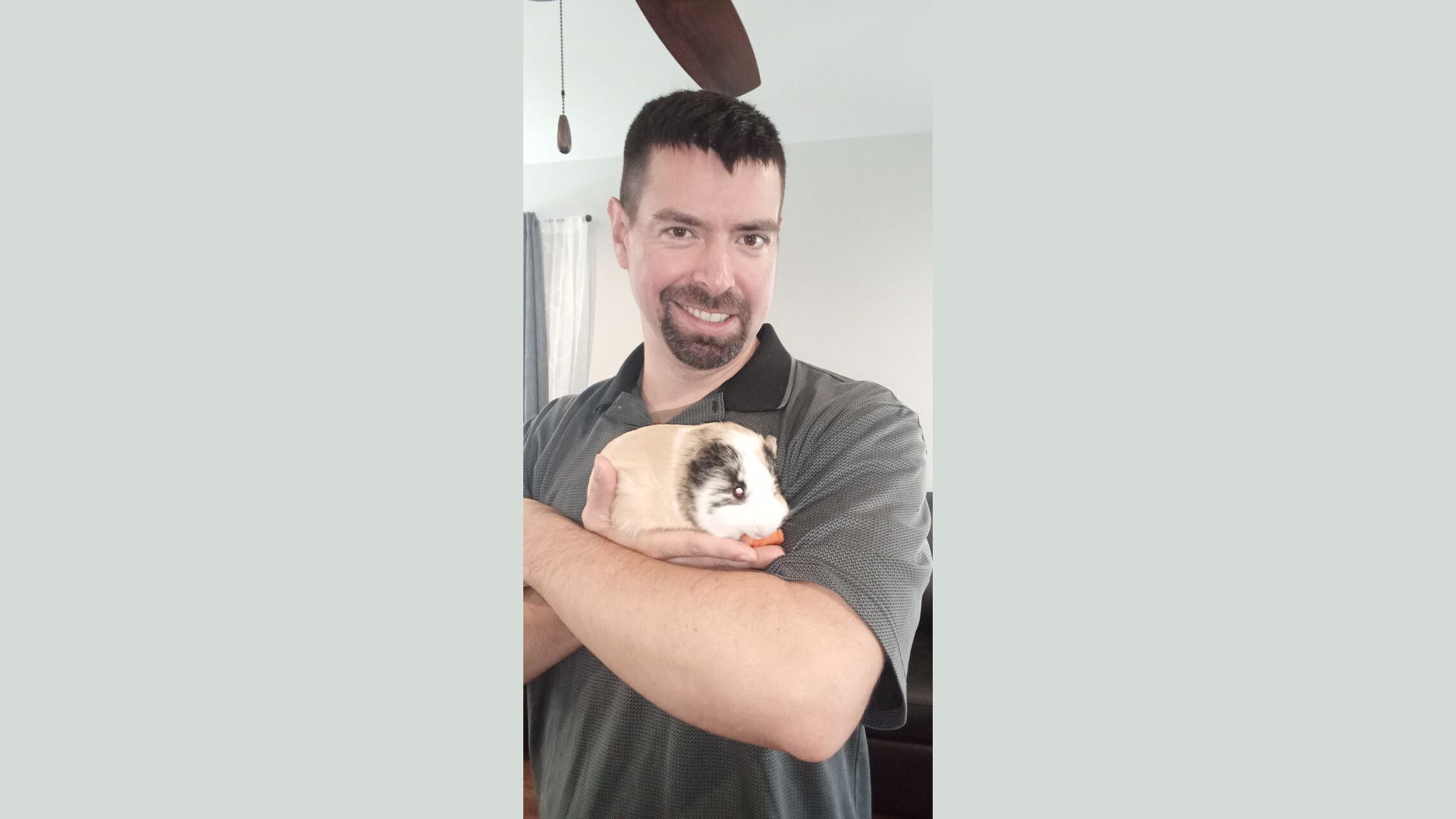 Man smiling while holding a guinea pig in his arms, indoors with a ceiling fan visible in the background.