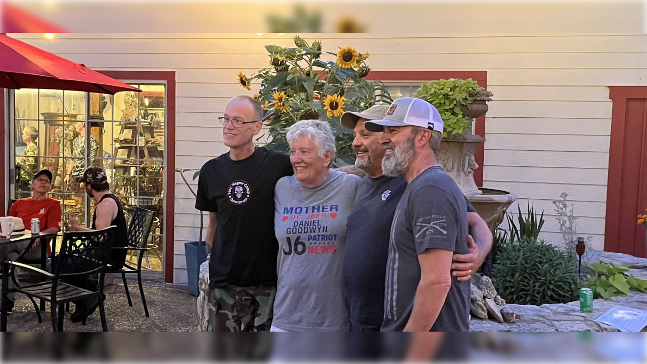 Group of four friends posing together outdoors, smiling in front of a garden with sunflowers and a decorative fountain.