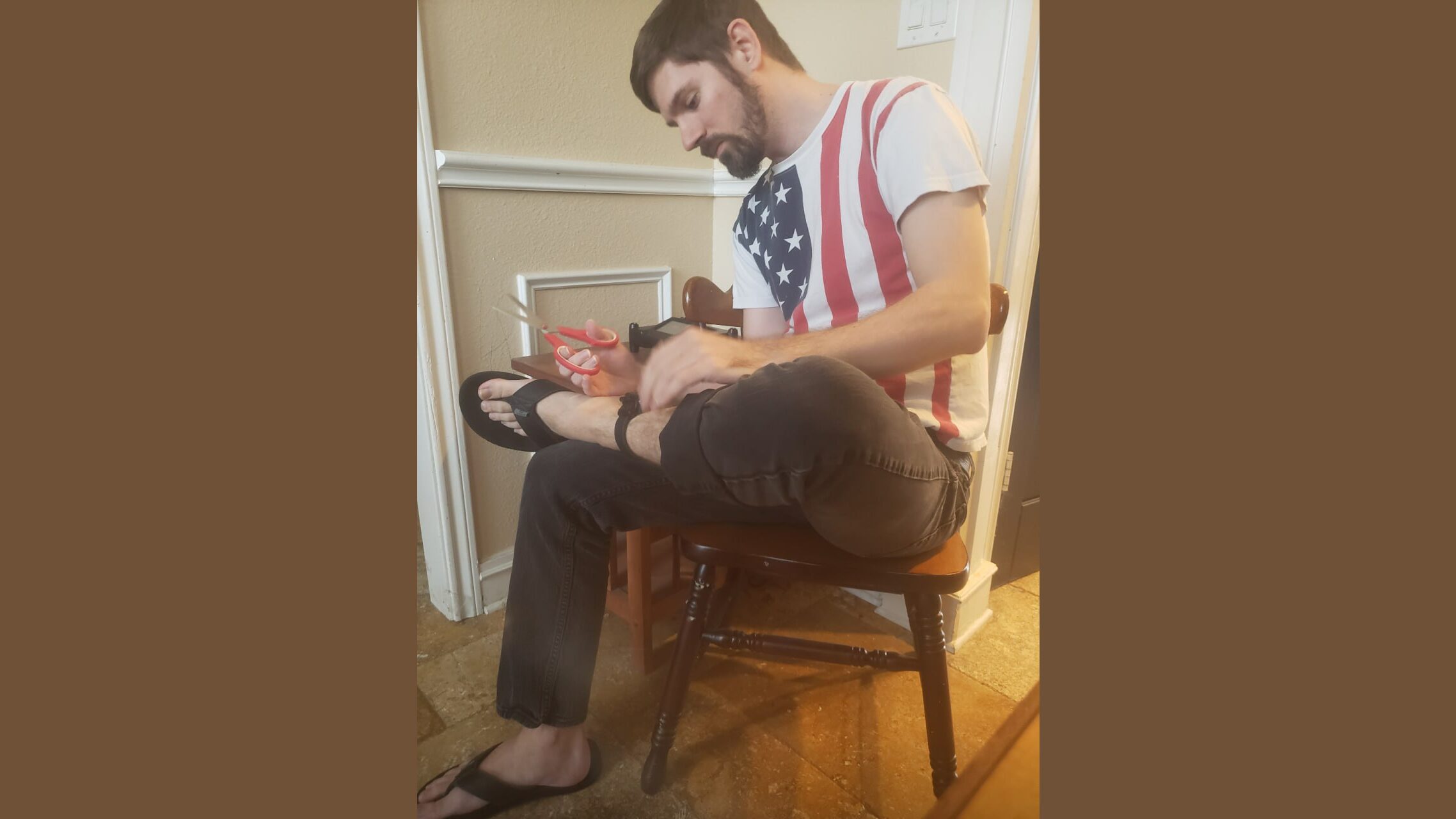 Man wearing an American flag t-shirt sits on a chair, cutting a piece of string while wearing flip-flops and black jeans.