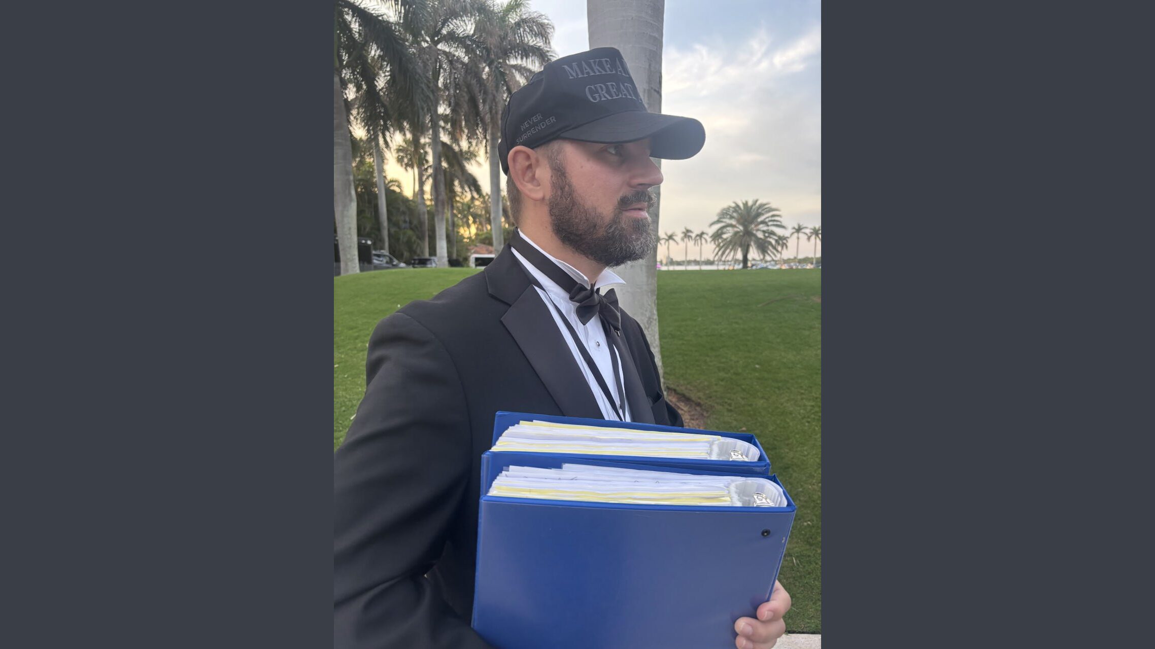 Man in a tuxedo and baseball cap holding blue binders, standing in a green park with palm trees in the background during sunset.