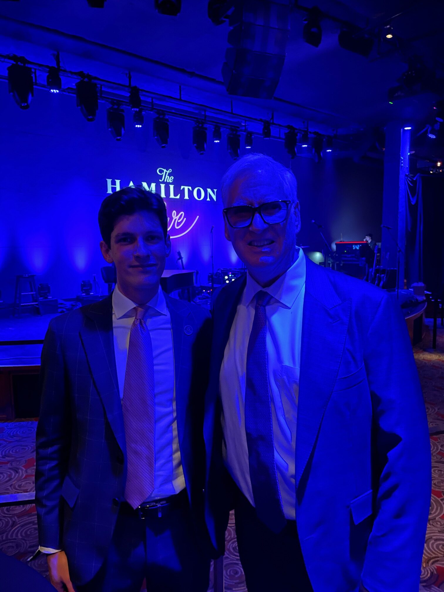 Two men pose together in front of a stage at The Hamilton Live, illuminated by blue lighting.
