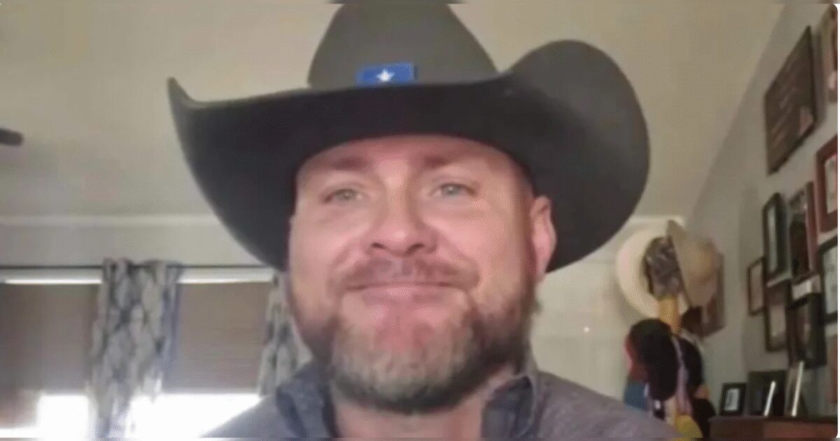 Man smiling in a cowboy hat, sitting indoors with decorative wall frames and hats in the background.