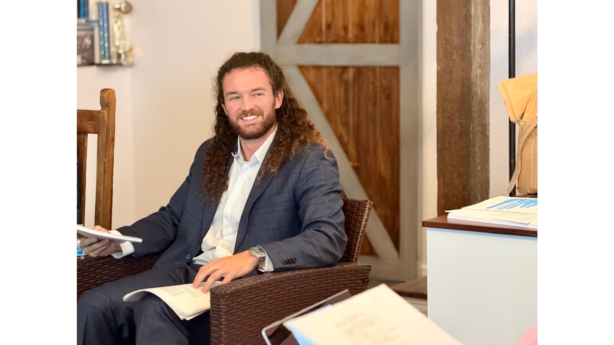 Man with curly hair and beard smiles while seated in a wicker chair, holding papers in a cozy indoor setting.