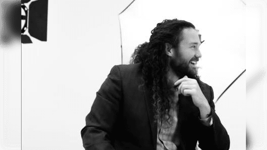 Man with curly hair and a beard smiling while seated, wearing a blazer, in a photography studio with soft lighting setups.