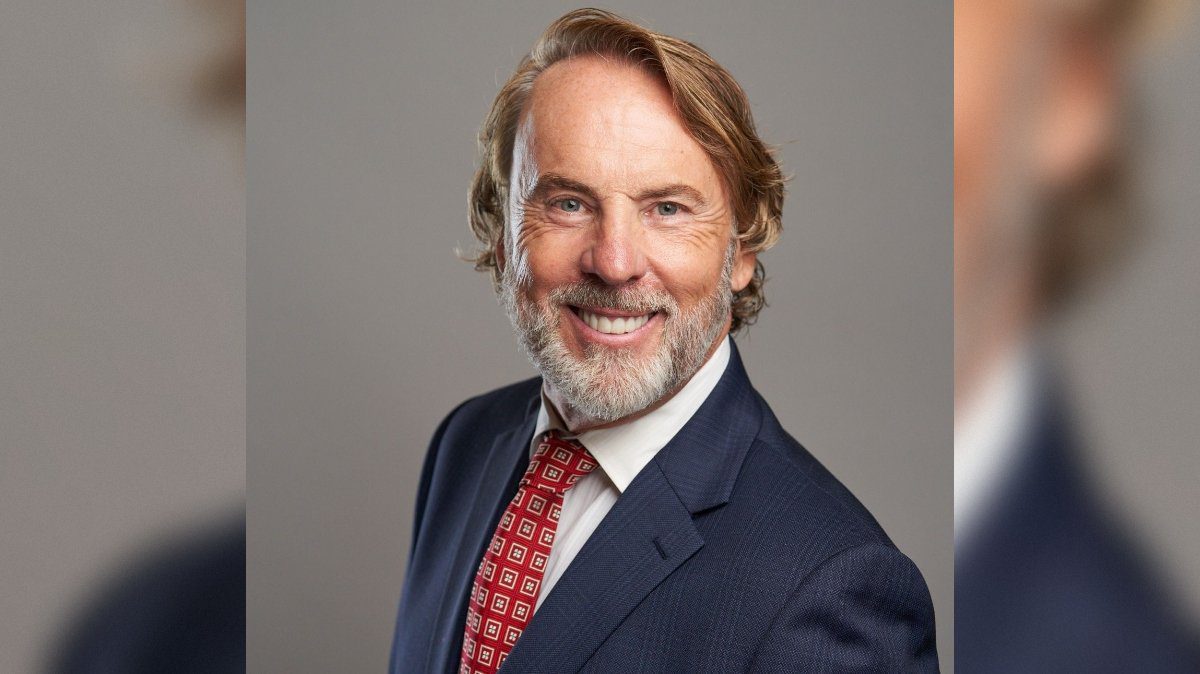 Professional headshot of a smiling man with a beard, wearing a navy suit and patterned red tie against a neutral background.