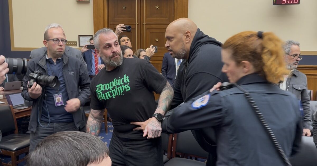 A tense moment during a congressional hearing, featuring a man in a black shirt being restrained by security while media capture the scene.