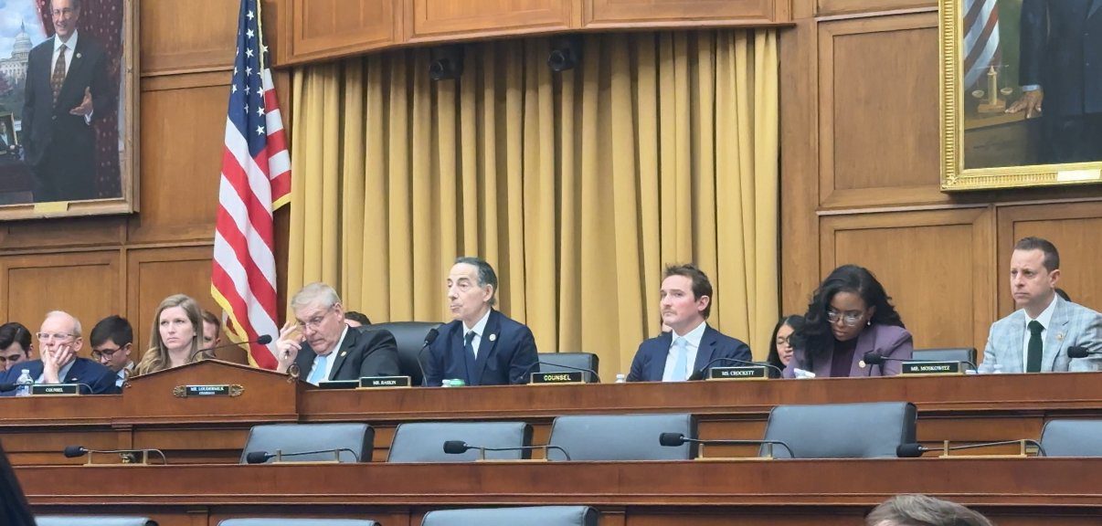 U.S. congressional hearing with several representatives seated at a wooden table, featuring an American flag and a portrait in the background.