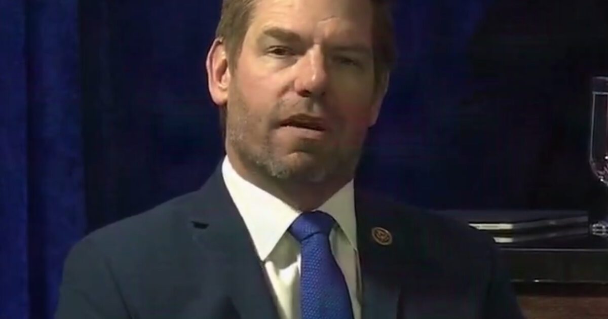 Man in a suit with a blue tie, seated against a blue backdrop, appearing thoughtful during a formal event.