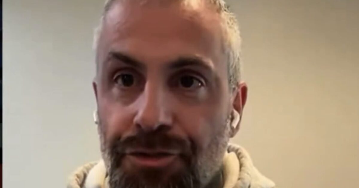 Man with a beard wearing a gray hoodie and earbuds, speaking in a well-lit indoor setting.