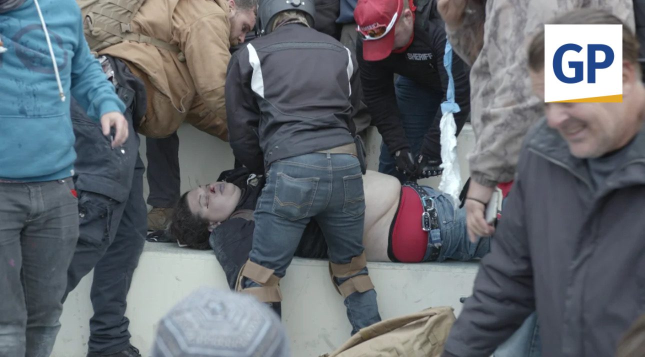 Emergency responders assist an injured person in a chaotic scene, with multiple individuals providing support and medical attention in a public space.