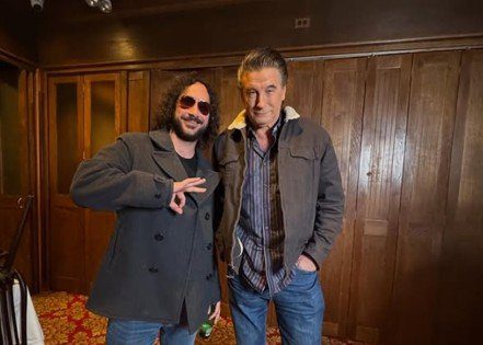 Two men posing together in a stylish indoor setting, one wearing sunglasses and a gray coat, the other in a casual shirt and jacket.