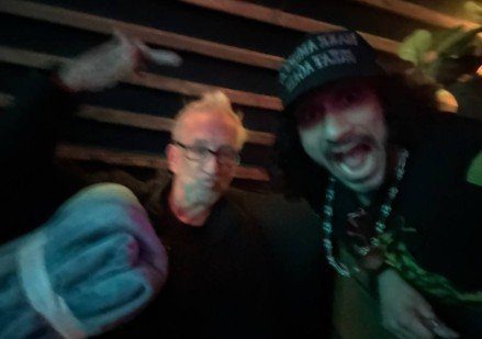 Two individuals posing for a photo in a dimly lit setting, one making a playful gesture while the other appears more serious.