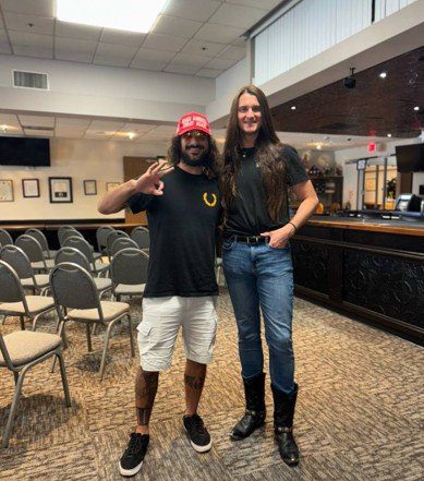 Two men pose together in a spacious indoor venue, one wearing a red cap and the other in a black shirt and cowboy boots, showcasing a casual friendship.