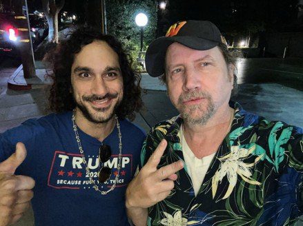 Two men posing for a selfie at night, one wearing a Trump-themed shirt and the other in a tropical print shirt, both smiling and making hand gestures.