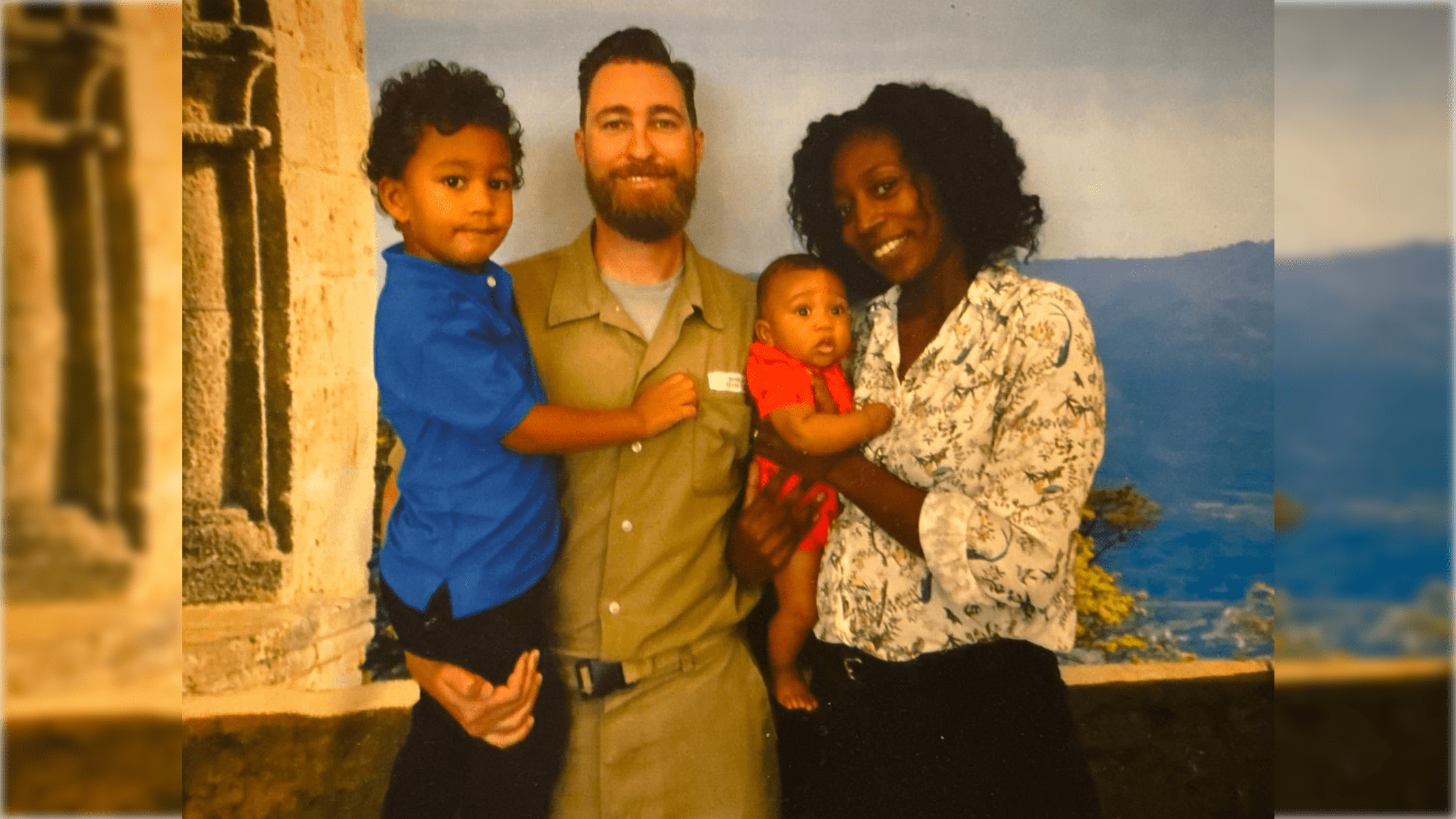 Family portrait featuring a smiling man in a khaki outfit holding a toddler, alongside a woman and an infant, set against a scenic backdrop.