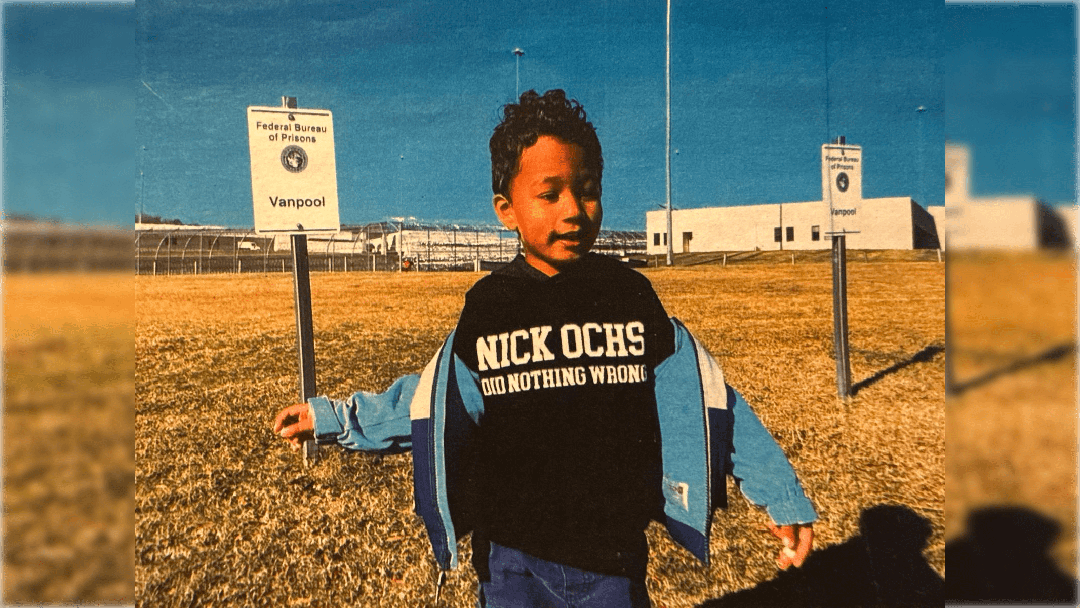 Young child in a black hoodie standing in front of a Federal Bureau of Prisons sign, with a grassy field and prison facility in the background.