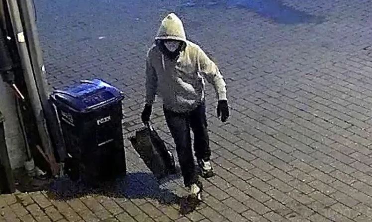 Individual in a gray hoodie and gloves carrying a bag near a trash can on a brick pavement at night.