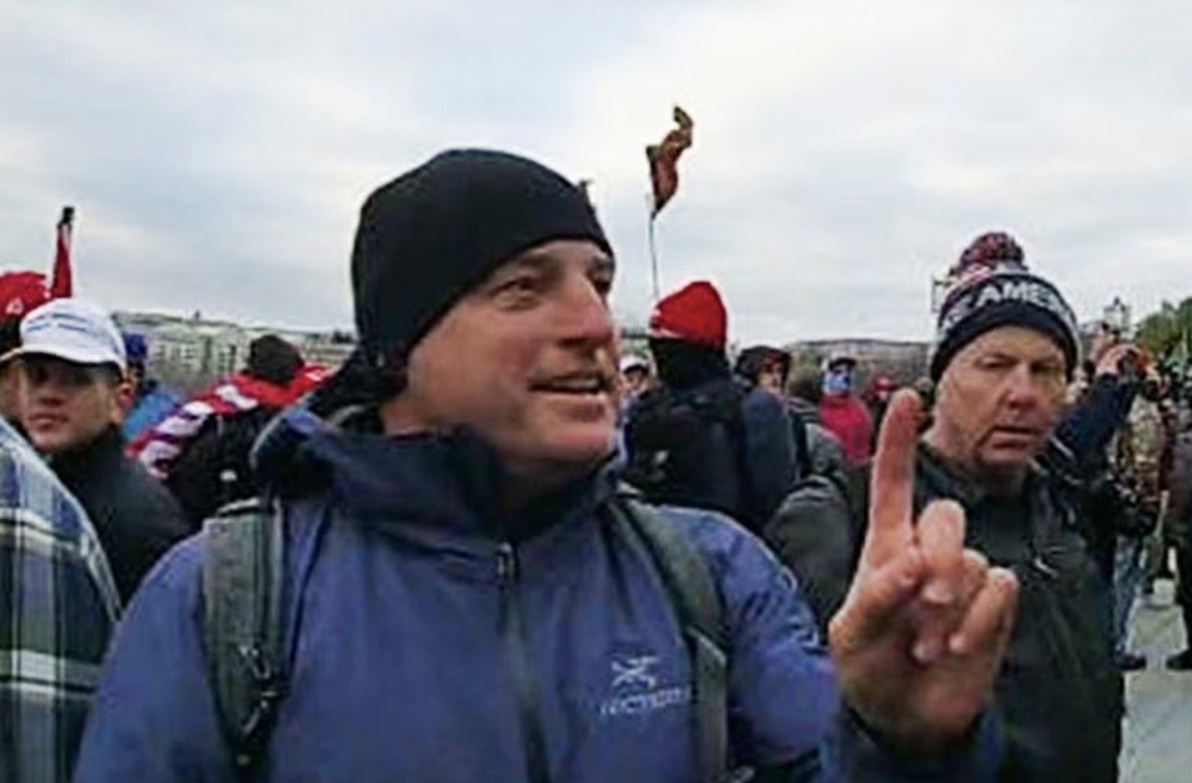 A man in a blue jacket and beanie gestures with one finger among a crowd at a rally, with flags and supporters visible in the background.