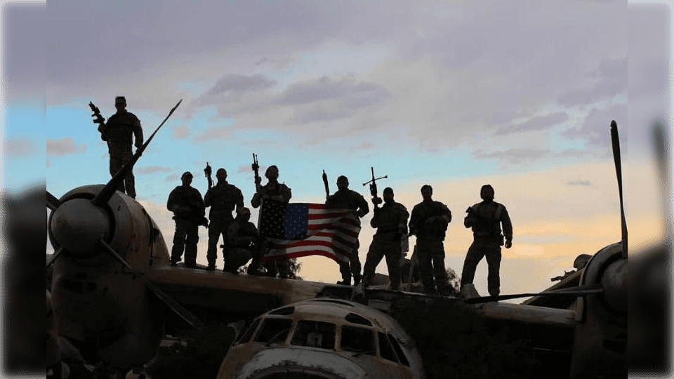 Silhouetted soldiers stand on a vintage aircraft holding an American flag against a dramatic sky, symbolizing patriotism and military service.