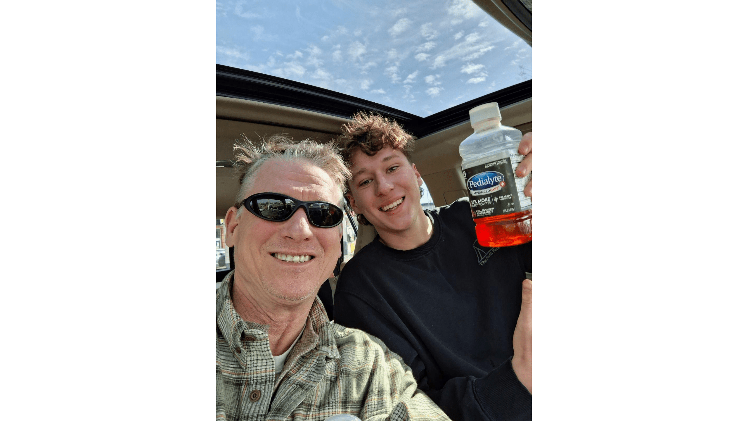 Two smiling men take a selfie in a car, one holding a bottle of Pedialyte, with a clear blue sky visible above.