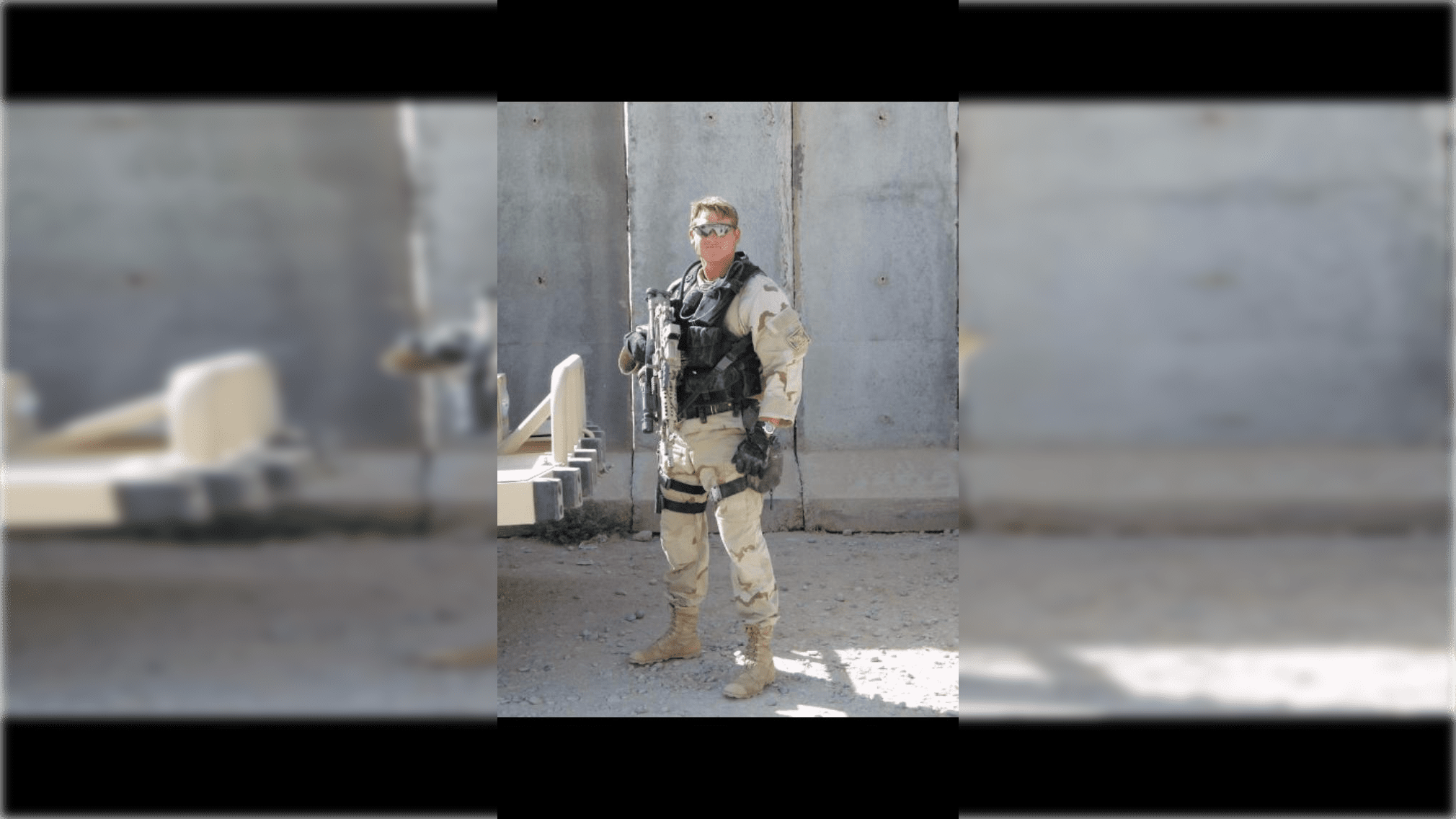Soldier in military uniform stands confidently with a firearm, set against a concrete backdrop in a desert environment.