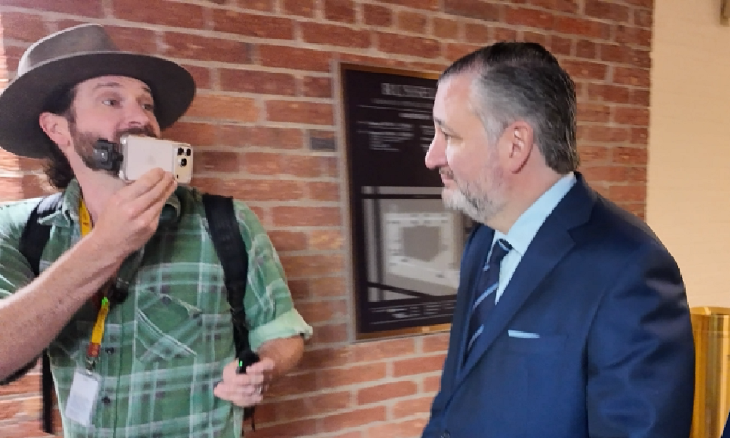 A man in a green plaid shirt and hat holds a phone while conversing with a suited politician in a brick hallway.