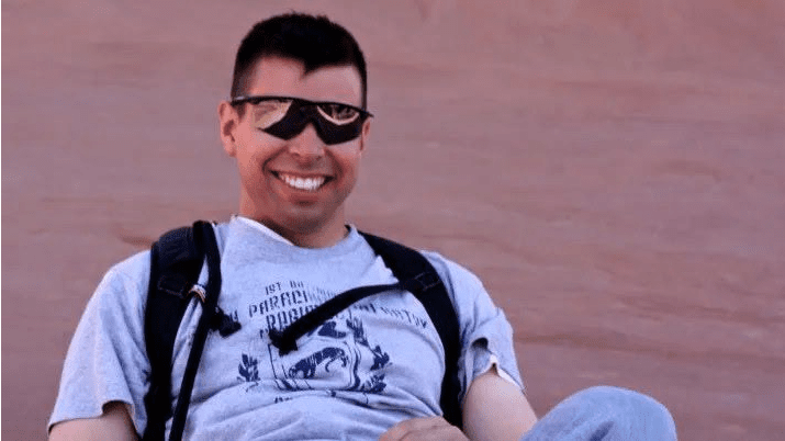 Smiling man wearing sunglasses and a gray t-shirt, sitting against a smooth, reddish-brown background, enjoying a relaxed moment outdoors.