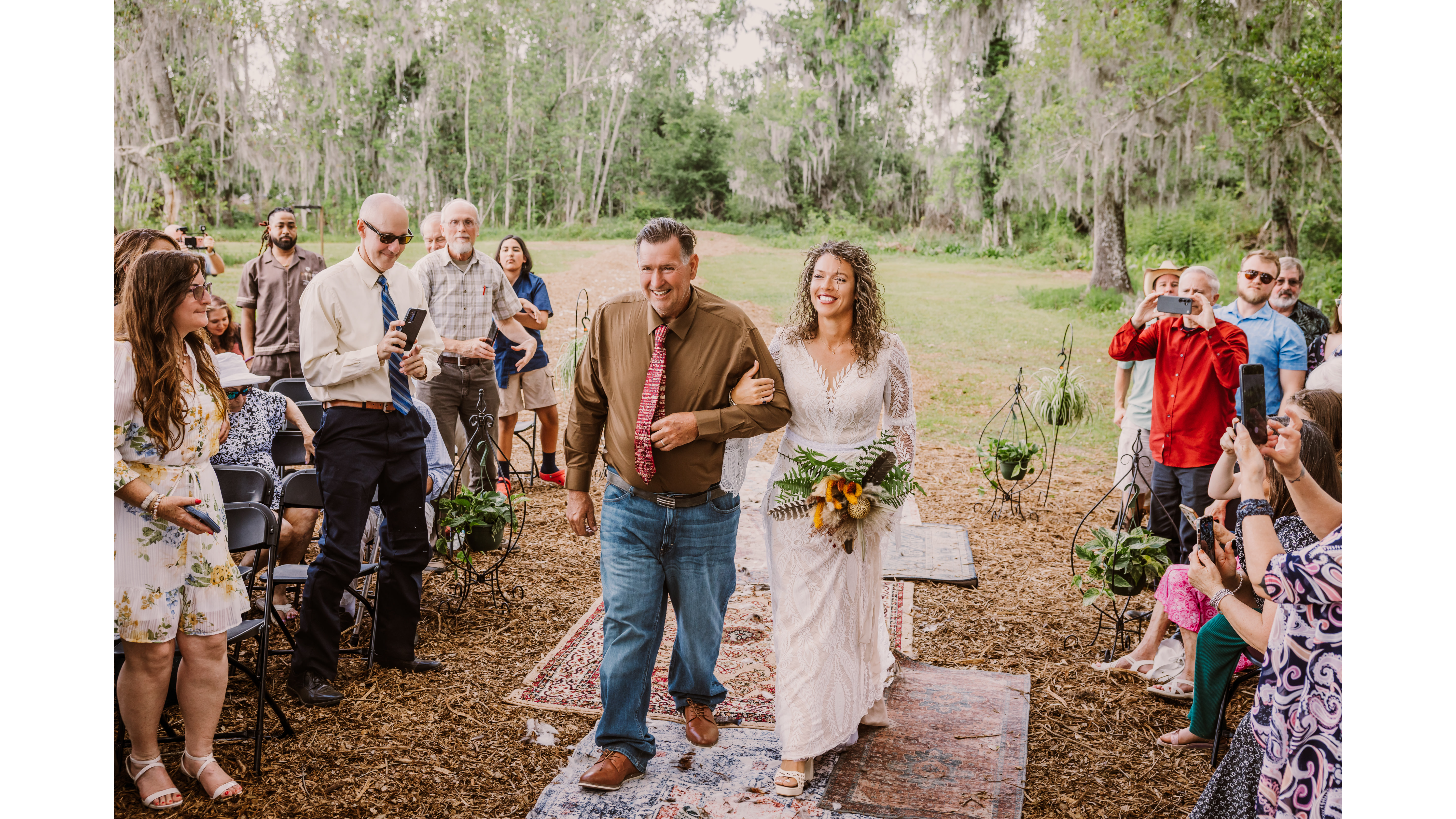 Bride walking down the aisle with her father at an outdoor wedding, surrounded by guests capturing the moment on their phones.
