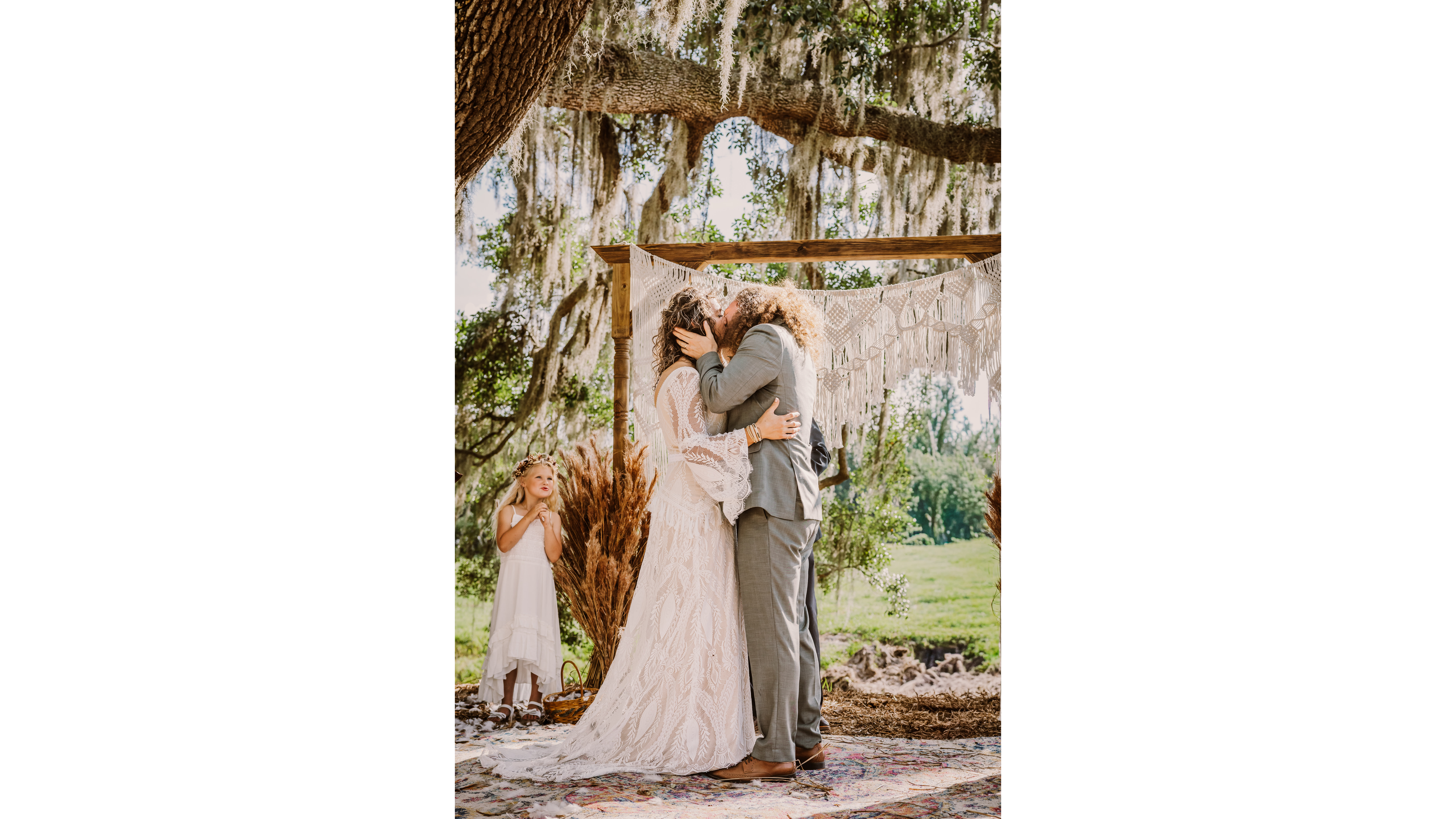 A couple shares a romantic kiss during their outdoor wedding ceremony, with a young girl in a white dress observing in the background.