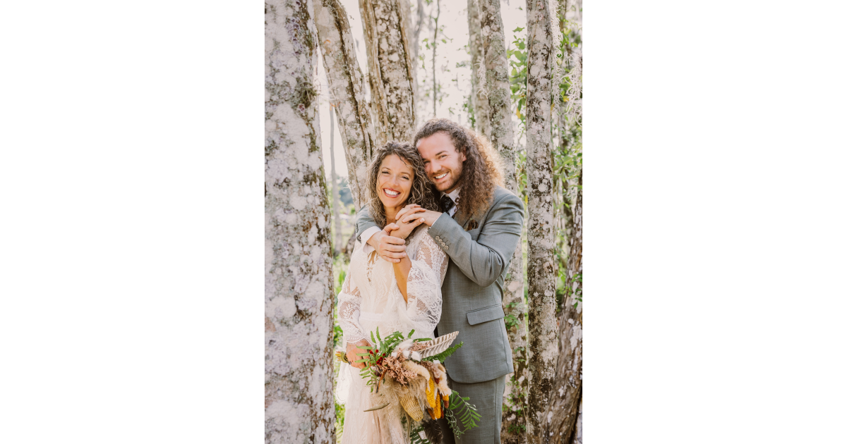 A joyful couple embraces in a forest setting, showcasing their wedding attire and a beautiful bouquet, with trees in the background.
