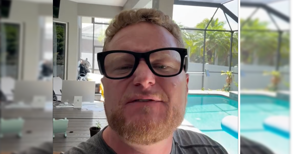 Man with glasses speaking in a bright room with a pool and greenery in the background, showcasing a relaxed home environment.