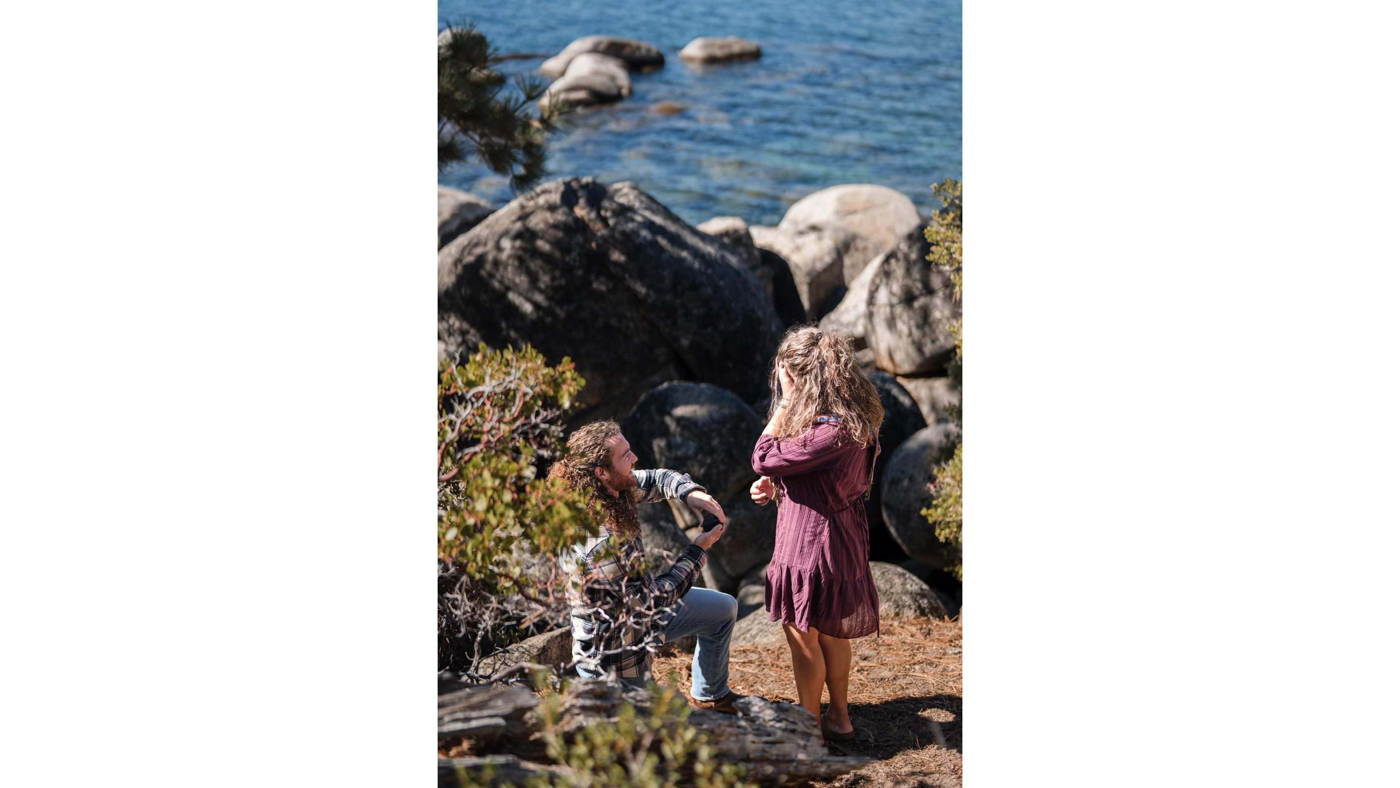 A man proposes to a woman by a serene lake surrounded by large rocks and greenery on a sunny day.